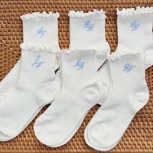 May include: Six pairs of white ankle socks with ruffled tops, each embroidered with light blue initials. The socks are arranged on a woven wicker surface. The socks are a ribbed knit.