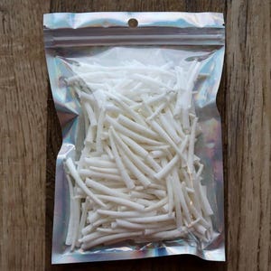Peut inclure: Un sac en plastique transparent et irisé rempli de nombreux objets cylindriques blancs. Le sac est scellé en haut et possède un petit trou pour l'accrochage. Le fond est une surface en bois.