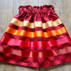 May include: A red plaid skirt with horizontal bands of gold, orange, and red ribbon. The skirt has a gathered waistband and a flared shape, creating a vibrant design. The skirt is made of a soft, flowing fabric.