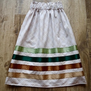 May include: A light-coloured skirt with a gathered waistband and a plaid pattern. The skirt is adorned with horizontal bands of green, brown, and gold ribbons. The skirt is made of fabric.