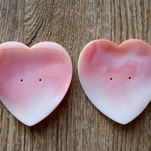 May include: Two heart-shaped soap dishes with a pink and white gradient. Each dish has two small holes. The dishes are on a wooden surface.