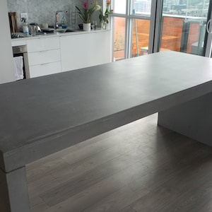 May include: A large, rectangular, grey concrete table with a smooth, polished surface. The table has a simple, modern design with a single, wide leg on each side.
