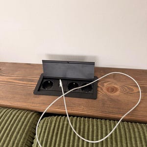 Op de afbeelding: Een zwarte pop-up stopcontact met drie stopcontacten en een oplaadkabel op een houten oppervlak. Het stopcontact is gedeeltelijk open en onthult de stopcontacten. De kabel is wit en aangesloten op een apparaat.