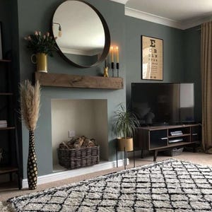 May include: A living room with a large round mirror, a wooden mantel, and a black and white patterned rug. A television sits on a dark wood console. The walls are painted a muted green.