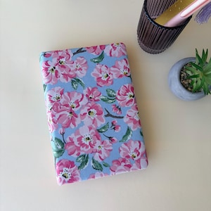 May include: A rectangular notebook featuring a light blue cover with a repeating pattern of pink flowers, green leaves, and brown stems. The notebook is positioned next to a dark purple glass cup containing a pink pen and a small potted succulent.
