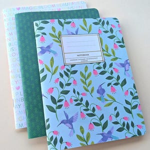May include: Three notebooks with floral and hummingbird designs. The front notebook has a light blue cover with a pattern of hummingbirds, pink flowers, and green leaves. The middle notebook has a green cover with a repeating pattern. The back notebook has a cover with text.