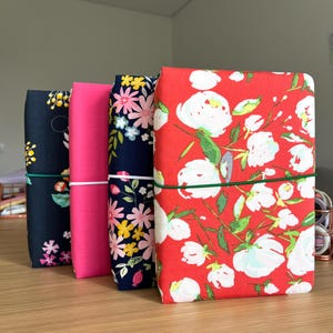 Puede incluir: Cuatro cuadernos cubiertos de tela con patrones florales. Los cuadernos están apilados uno encima del otro. El cuaderno superior es rojo con flores blancas, el segundo cuaderno es azul marino con flores amarillas y rosas, el tercer cuaderno es rosa con un patrón de lunares blancos y el cuaderno inferior es azul marino con lunares blancos y grises.