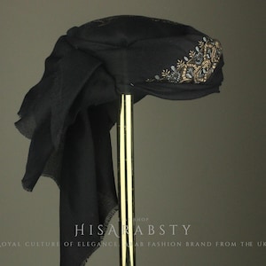 Può includere: Un turbante nero con ricami floreali dorati, blu e bianchi. Il turbante è drappeggiato e ha una lunga sciarpa fluente. Il testo "HISAR ABSTY" e "ROYAL CULTURE OF ELEGANCE" è visibile.