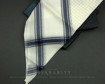 May include: A white and blue patterned shemagh, a traditional Arab headscarf, folded and displayed on a black surface. The shemagh has a checkered pattern with blue stripes and a white background.