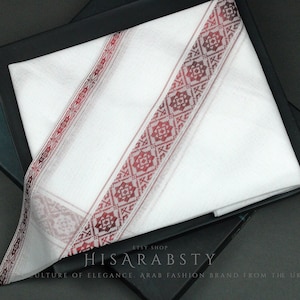 Puede incluir: Keffiyeh de algodón blanco con un borde bordado en rojo y blanco. El keffiyeh está doblado y se muestra en una caja negra. La caja tiene el texto "HISARABSTY CULTURE OF ELEGANCE ARAB FASHION BRAND FROM THE UK" impreso en ella.