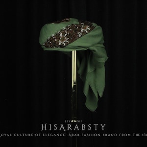 May include: A sage green head wrap featuring a wide, dark brown band adorned with intricate metallic beadwork and embroidery. The fabric is draped with a soft, flowing tail hanging down one side, displayed on a vertical stand with text below.