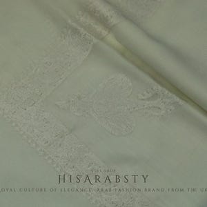 May include: Cream-coloured fabric with delicate lace edging and an embroidered motif. The fabric is folded, creating shadows and texture. The text "HISARABSTY" and "ROYAL CULTURE OF ELEGANCE - ARAB FASHION BRAND FROM THE UK" are visible.