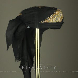 May include: A black turban with gold embroidery on the front. The turban is draped over a gold stand. The text "HISAR ABSTY ROYAL CULTURE OF ELEGANCE AB FASHION BRAND FROM THE UK" is at the bottom of the image.