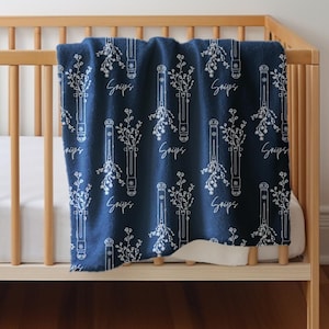 Puede incluir: Una cuna de madera con un colchón blanco y una manta de patrón azul y blanco. La manta tiene un diseño floral y las palabras "Snugs" repetidas en toda la superficie.