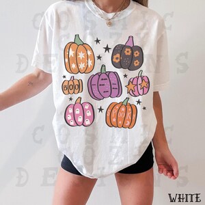 May include: White t-shirt with a Halloween design. The graphic features pumpkins in orange, purple, and pink, decorated with stars and the words "BOO" and "SPOOKY".