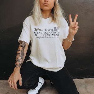 May include: Cream-colored t-shirt with the text "THE TORTURED TATTOO ARTISTS DEPARTMENT" and the phrase "all's fair in love and cover ups." The shirt features a tattoo gun graphic. The person is wearing black jeans and white shoes.