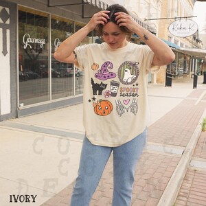 May include: Ivory-colored t-shirt with a Halloween-themed graphic. The design includes a witch's hat, a black cat, a pumpkin, a ghost, and the words "Spooky Season". The shirt is a comfortable, casual style.