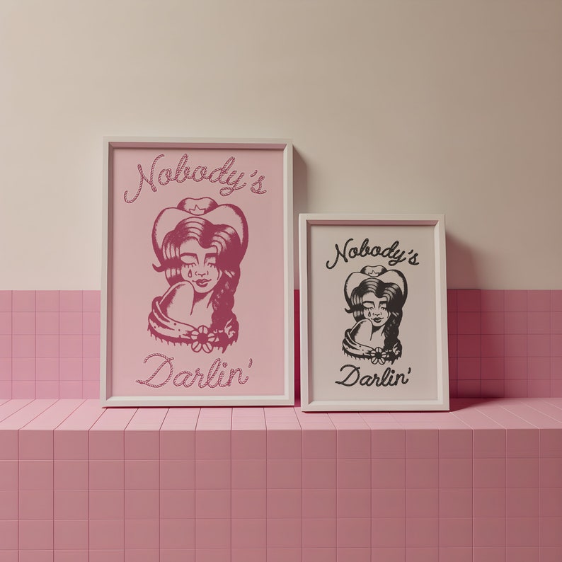 May include: Two framed prints with the text "Nobody's Darlin'" and a black and white illustration of a woman wearing a cowboy hat. The larger print has a pink background and the smaller print has a white background.