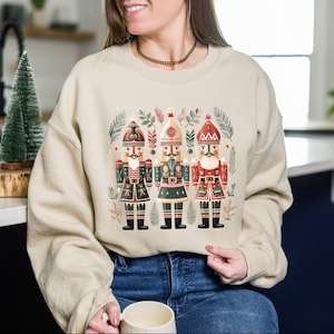 May include: Beige sweatshirt featuring a festive design of three nutcrackers in red, green, and black uniforms. The nutcrackers are surrounded by botanical elements. The sweatshirt is a comfortable, casual style.