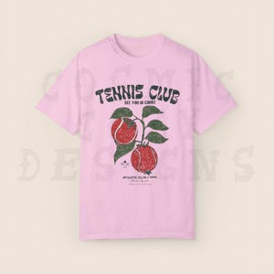 May include: Pink t-shirt with a vintage tennis club graphic. The graphic features a tennis racket, two tennis balls, and the text "Tennis Club See You In Court Athletic Club / 1990"