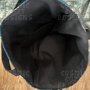 Pu&ograve; includere: Vista aperta di una borsa tote con interno nero ed esterno fantasia. La borsa ha un cinturino nero e le parole "COSMIC COZY DESIGNS" sono visibili sul lato.