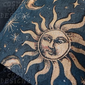 May include: A decorative pillow with a celestial design. The pillow features a detailed illustration of a sun with a face, surrounded by stars and crescent moons, set against a dark blue background. The pillow has a textured appearance.