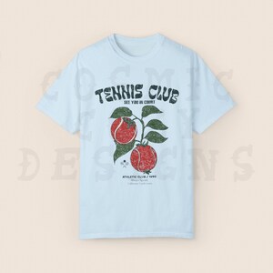 May include: Light blue t-shirt with a vintage-style graphic of a tennis club logo. The logo features the text "Tennis Club" with the words "See you in court!" underneath. The logo is surrounded by a graphic of two red tennis balls on a vine.