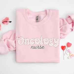 Könnte beinhalten: Rosafarbener Pullover mit dem Text "Oncology nurse" in einem Muster aus weißen und rosa Schleifen.