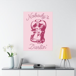 May include: Pink background with a pink illustration of a woman wearing a cowboy hat and a flower in her hair. The text "Nobody's Darlin'" is written in a pink rope-like font.