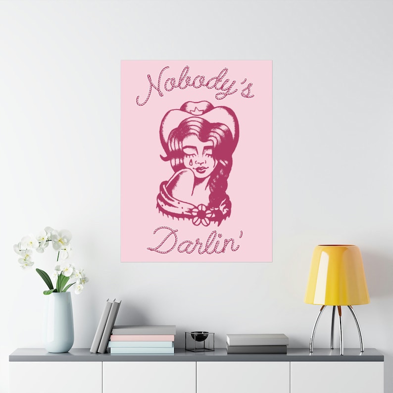 May include: Pink background with a pink illustration of a woman wearing a cowboy hat and a flower in her hair. The text "Nobody's Darlin'" is written in a pink rope-like font.
