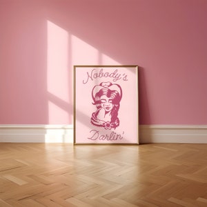 May include: A pink framed print with a pink illustration of a woman wearing a cowboy hat and the text "Nobody's Darlin'" in a pink rope-like font.