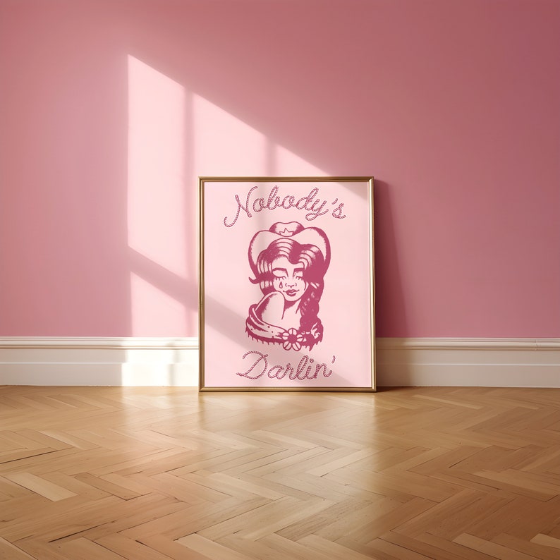 May include: A pink framed print with a pink illustration of a woman wearing a cowboy hat and the text "Nobody's Darlin'" in a pink rope-like font.