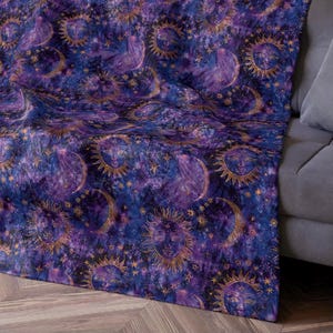 May include: A purple and gold throw blanket with a repeating pattern of suns and moons. The suns have faces and are surrounded by stars.