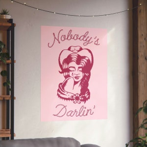 May include: Pink poster featuring a red illustration of a woman wearing a cowboy hat and a flower on her shoulder. The text "Nobody's Darlin'" is written in a rope-like font.