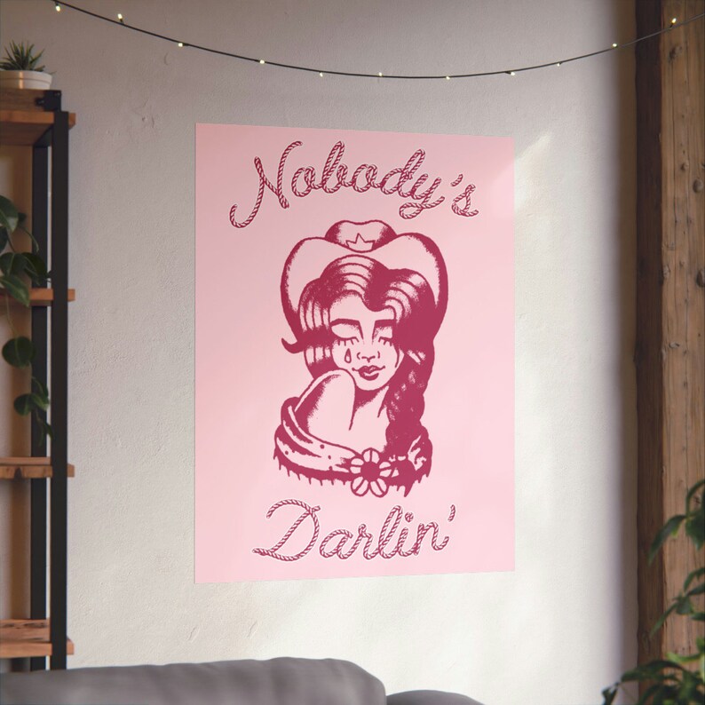 May include: Pink poster featuring a red illustration of a woman wearing a cowboy hat and a flower on her shoulder. The text "Nobody's Darlin'" is written in a rope-like font.