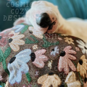 May include: A white bull terrier dog with a black nose is sleeping on a brown floral patterned pillow. The pillow has pink, blue, and yellow flowers.