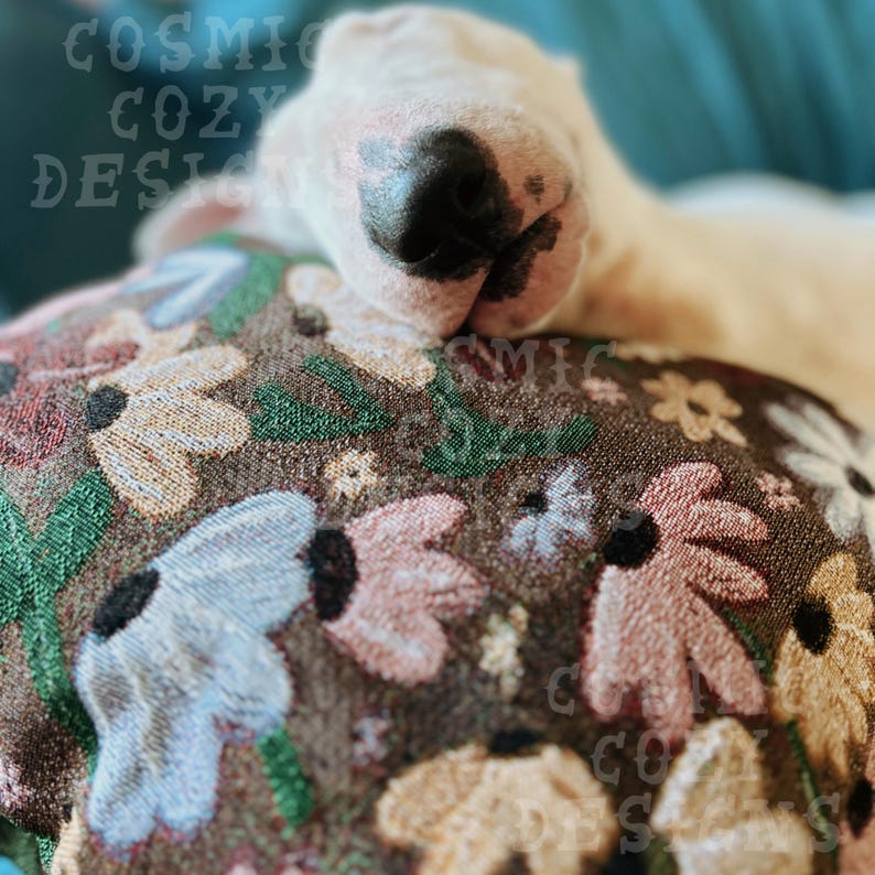 May include: A white bull terrier dog with a black nose is sleeping on a brown floral patterned pillow. The pillow has pink, blue, and yellow flowers.
