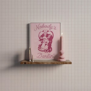 May include: A pink framed print with a pink illustration of a woman wearing a cowboy hat. The text on the print reads "Nobody's Darlin'". Two white candles are on a wooden shelf below the print.