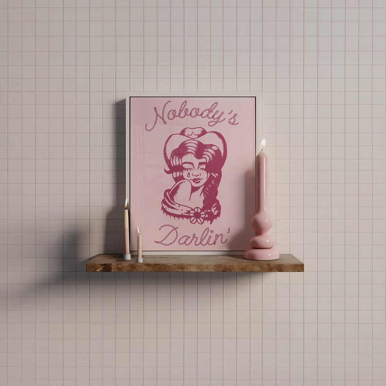 May include: A pink framed print with a pink illustration of a woman wearing a cowboy hat. The text on the print reads "Nobody's Darlin'". Two white candles are on a wooden shelf below the print.
