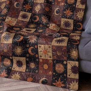 以下が含まれることがあります： パッチワークデザインの、天体モチーフのスローケット。茶色、金色、黒の色合いで太陽、月、星が描かれた正方形が特徴です。グレーのソファに掛けられています。