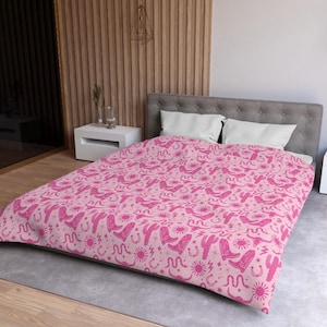 May include: A pink duvet cover with a repeating pattern of pink cowboy boots, cacti, suns, horseshoes, and snakes. The duvet cover is on a bed with white pillows and a wooden headboard.