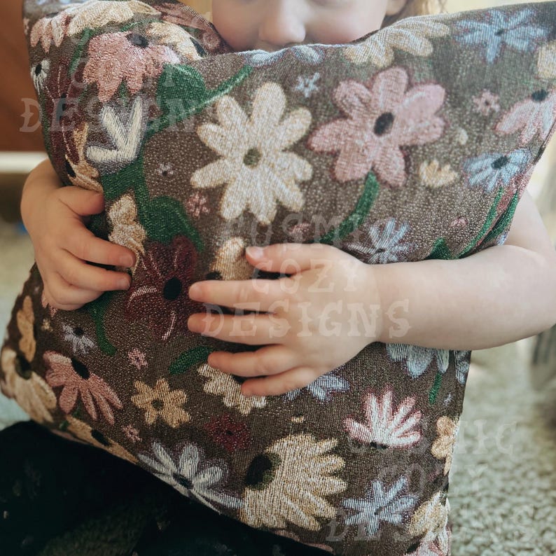 May include: A child holds a brown throw pillow with a floral pattern. The pillow features embroidered flowers in shades of pink, white, and green.