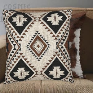 Puede incluir: Cojín decorativo con un patrón geométrico en negro, marrón y crema. El cojín presenta un diseño de diamantes y triángulos, con un segundo cojín con un patrón de piel de vaca visible en el fondo. La palabra "COSMIC" es visible en la esquina superior izquierda.