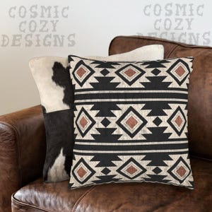Puede incluir: Dos cojines decorativos sobre un sofá de cuero marrón. Uno presenta un patrón de piel de vaca en blanco y negro. El otro tiene un diseño geométrico en negro, beige y marrón. El texto "COSMIC COZY DESIGNS" es visible en el fondo.