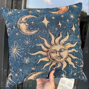 May include: Square decorative cushion with a celestial design. The cushion features a dark blue background with a pattern of the sun, moon, and stars in shades of gold and cream. The cushion is approximately 40 cm square.