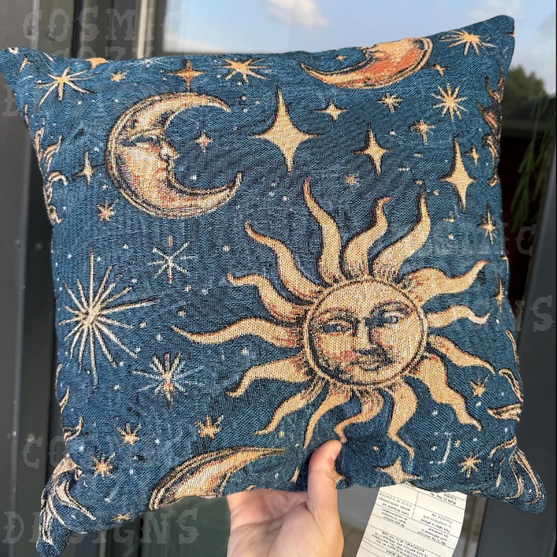 May include: Square decorative pillow with a celestial design. The pillow features a dark blue background with a pattern of the sun, moon, and stars in shades of gold and cream. The pillow is approximately 16 inches square.