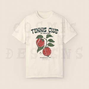 May include: White t-shirt with a vintage-style graphic of tennis rackets and tennis balls. The text on the shirt reads "Tennis Club See You In Court Athletic Club / 1990"