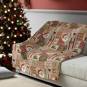 Pode incluir: Uma manta de Natal com um design de patchwork com o Pai Natal e árvores de Natal. A manta, em tons de vermelho, verde e creme, está sobre um sofá. Uma árvore de Natal decorada é visível no fundo.