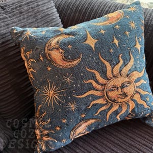 May include: Square decorative pillow with a celestial design. The pillow features a dark blue background with gold and tan suns, moons, and stars. The pillow is made of a textured fabric and is placed on a dark blue corduroy surface.