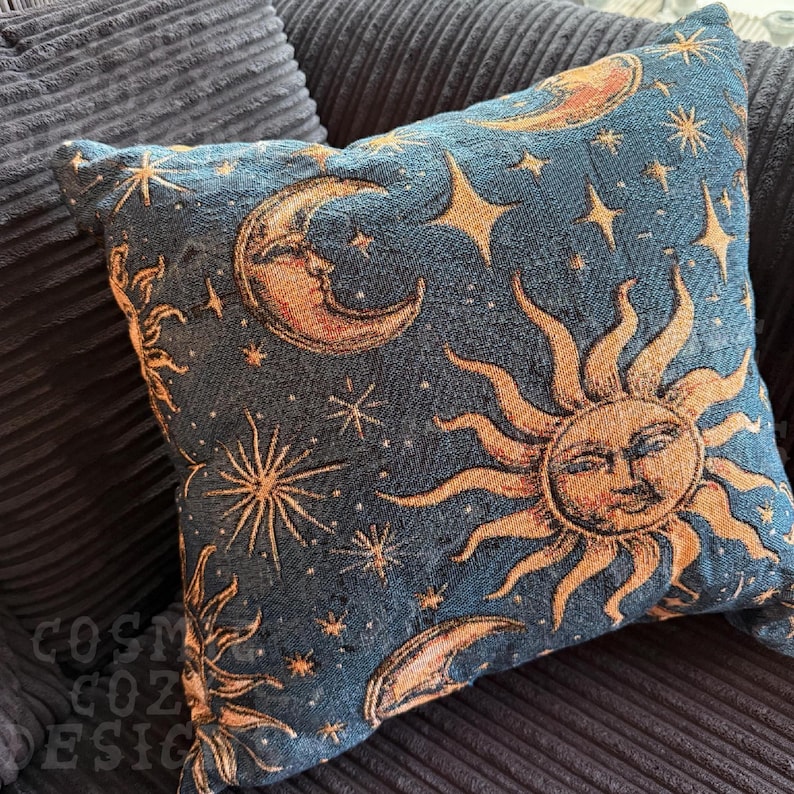 May include: Square decorative pillow with a celestial design. The pillow features a dark blue background with gold and tan suns, moons, and stars. The pillow is made of a textured fabric and is placed on a dark blue corduroy surface.